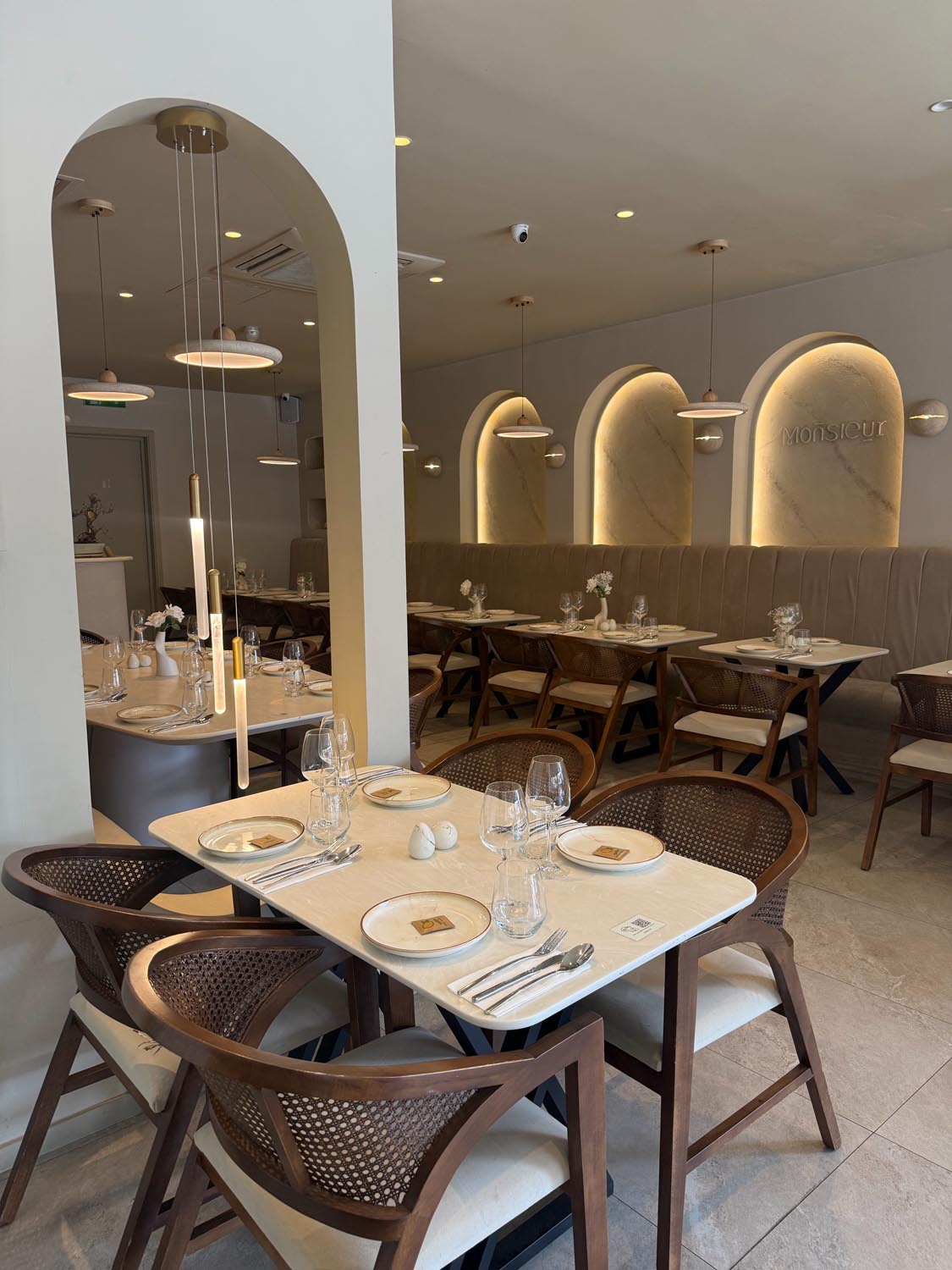 Arched mirror reflecting pendant lighting beside laid tables with cane-backed walnut chairs